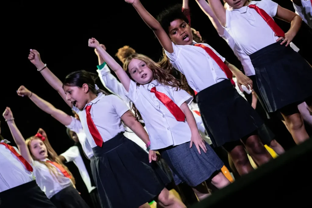 Junior performers from Cherry Stars Stage and Screen School dressed in school uniforms with red ties, singing and gesturing during the production at Stanley Arts.